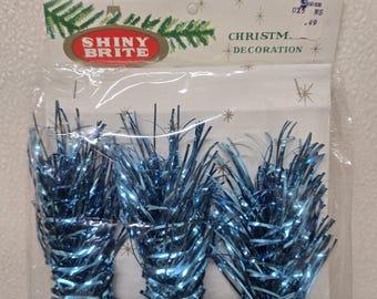 May include: Vintage Shiny Brite Christmas decoration. The package contains three bundles of blue tinsel. The packaging has the words "Shiny Brite" in a red oval and "Christmas Decoration" printed on the top.