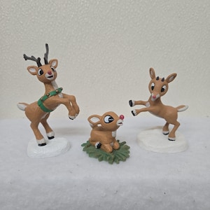 May include: Three brown reindeer figurines. One is leaping with antlers, wearing a green harness. Another is seated on green foliage, and the third is mid-jump. All have red noses and are on white bases.