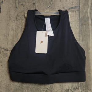 May include: A black sports bra with a scoop neck and a wide band at the bottom. A white tag with the brand logo and product information hangs from the bra. The bra is hanging on a clear plastic hanger against a wooden background.