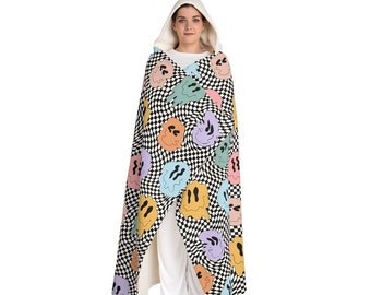 May include: A hooded blanket featuring a black and white checkered pattern with colorful, melting smiley face designs. The blanket has a white hood and is worn by a person.