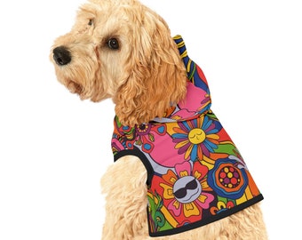 May include: A light brown dog wearing a colorful hooded dog vest with a psychedelic pattern of flowers and smiley faces. The vest has a black trim.