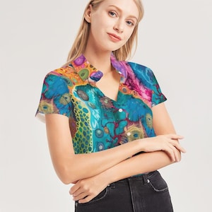 Puede incluir: Una camisa de manga corta con un vibrante estampado abstracto en tonos de azul, turquesa, rosa, naranja y amarillo. La camisa tiene cuello y botones blancos. El diseño general es artístico y colorido.