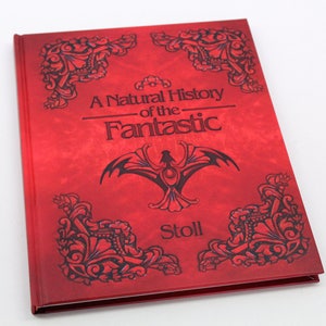 May include: A hardcover book titled "A Natural History of the Fantastic" with a red cover. The cover features black ornate designs and a central emblem. The author's name, Stoll, is printed at the bottom.
