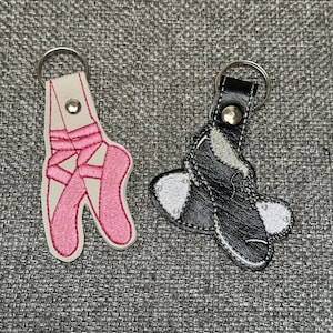 May include: Two keychains featuring embroidered designs. One keychain is cream-coloured with a pink ballet slipper motif. The other keychain is black with a white tap shoe motif. Both items include a silver-toned metal ring and a snap button closure.