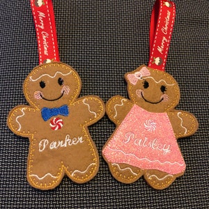 May include: Two gingerbread person ornaments with red ribbon hangers. The gingerbread person on the left is brown with a blue bow tie and the name "Parker" embroidered on it. The gingerbread person on the right is pink with a pink bow and the name "Paisley" embroidered on it.