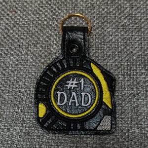May include: Black leather keychain with a gold ring. The keychain has a circular design with the text "#1 DAD" in white lettering, surrounded by a yellow border. A thoughtful gift.