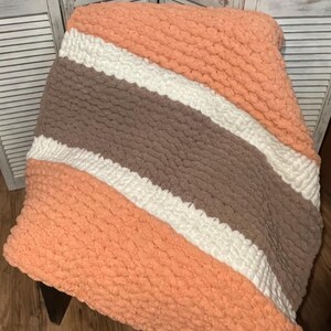 Può includere: Una morbida coperta a maglia grossa a righe pesca, bianche e marroni. La coperta è piegata, mostrando il suo aspetto strutturato e accogliente. Questa coperta fatta a mano è perfetta per aggiungere calore e stile a qualsiasi stanza.