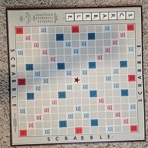 May include: A Scrabble board game with a grid of squares in various colors, including red, blue, and pink. The board has the word "SCRABBLE" along the sides. The game board is rectangular with a brown border and a star in the center.