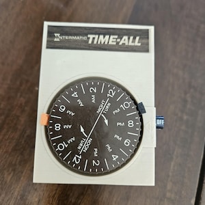 Peut inclure: Un minuteur blanc Intermatic TIME-ALL avec un cadran noir. Le cadran affiche les heures AM et PM, avec un pointeur indiquant l'heure actuelle. Le minuteur a un levier orange et un interrupteur OFF bleu.