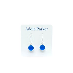 May include: A pair of Addie Parker earrings. Each earring features a small, round, bright blue charm suspended from a silver-colored hook. The earrings are displayed on a white card with the brand name printed in blue.