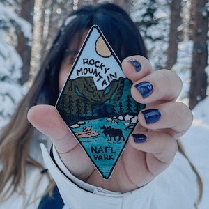 May include: A person in a white jacket holds up a diamond-shaped patch with a blue and green illustration of a mountain range, a lake, and a moose. The patch reads "Rocky Mountain Natl Park".