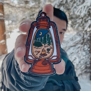 May include: A red and white lantern patch featuring a camping scene. The scene depicts a campfire, a tent, a mountain, and a crescent moon. The patch is held against a snowy forest backdrop.