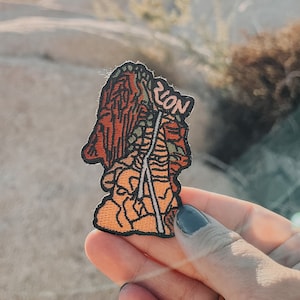 Op de afbeelding: Een bruin en oranje geborduurde patch met een rotsformatie en het woord "Zion" erop. De patch wordt in een hand gehouden tegen een achtergrond van grote rotsen.