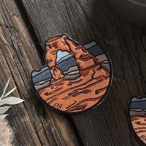 May include: Three embroidered patches featuring the Delicate Arch in Arches National Park, Utah. The patches are brown, orange, and gray with a blue sky and white clouds in the background.