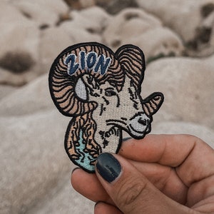 Op de afbeelding: Een geborduurde patch van een dikhoornschaapskop in bruin en wit, met het woord "ZION" in blauw geborduurd op de bovenkant van de kop. De patch wordt in een hand gehouden.