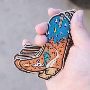 Könnte beinhalten: Ein Stapel gestickter Bügelbilder mit einem Cowboy-Stiefel, in dem sich eine Wüstenlandschaft befindet. Der Stiefel ist blau mit weißen Sternen und einem Halbmond. Die Wüstenlandschaft ist orange mit einem Kaktus, einer Straße und einem Totenkopf.