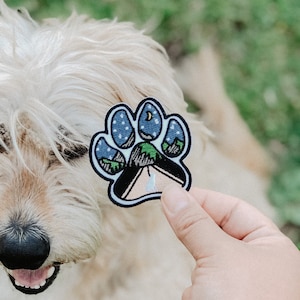 Könnte beinhalten: Ein weißer Hund mit schwarzer Nase schaut auf einen Pfotenabdruck-Patch mit einer Camping-Szene, einem Mond und Sternen. Der Patch wird in einer Hand gehalten.