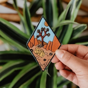 Puede incluir: Un parche en forma de diamante con un paisaje desértico con un árbol de Josué, montañas y un sol. El parche tiene las palabras "CAMP LIFE" y se sostiene sobre un fondo de follaje verde y marrón.
