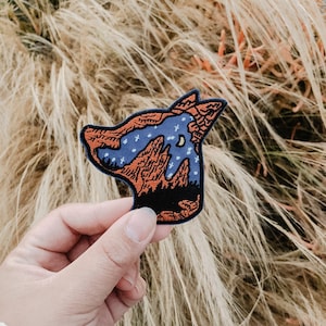 Pode incluir: Um remendo bordado azul e laranja de uma cabeça de lobo com uma cordilheira e uma lua dentro. O remendo está sendo segurado por uma mão.