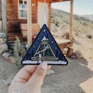 May include: A blue triangular patch featuring an embroidered image of a small cabin with a Christmas tree inside. The cabin has a white roof and a yellow door. The cabin is surrounded by snow and stars.