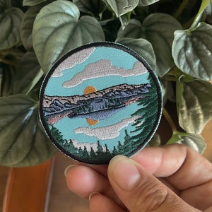 Pode incluir: Um remendo bordado redondo com uma cena de lago azul. O lago é cercado por árvores verdes e montanhas. O céu é azul com nuvens brancas e um sol amarelo.