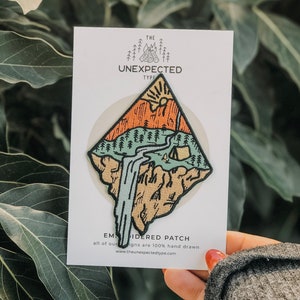 May include: A brown, orange, and green embroidered patch with a mountain scene, a waterfall, and a tent. The patch is on a white card with the text "THE UNEXPECTED TYPE" and "EMBROIDERED PATCH" in black.