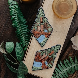 May include: Two embroidered patches featuring a forest scene with tall trees and a winding river. The patches are diamond-shaped and have a rustic brown and green color scheme.