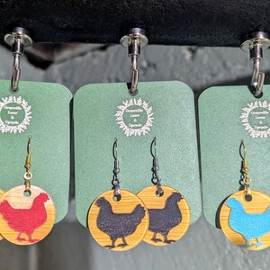 May include: Three pairs of earrings with a chicken design. Each pair features a circular, light-colored wooden base with a chicken silhouette in red, black, or blue. The earrings hang from green tags with the text "Greensville Laser & Upcycle".