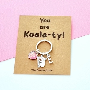 May include: A brown card with the text "You are Koala-ty!" and a silver keyring with a pink glitter heart, a silver koala charm, and a silver letter "E" charm. The card is on a pink background.