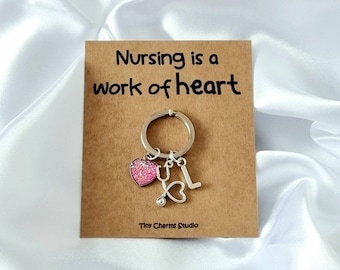 May include: A silver keychain with a pink glitter heart charm, a stethoscope charm, and a letter L charm. The keychain is on a brown card with the text "Nursing is a work of heart" and "Tiny Charms Studio".