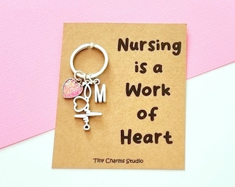 May include: A silver keychain with a pink glitter heart charm, a silver letter "M" charm, and a silver medical symbol charm. The keychain is attached to a brown card with the text "Nursing is a work of heart. Tiny Charms Studio."