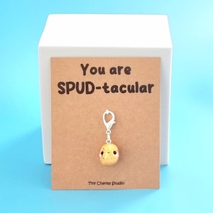 May include: A brown card with the text "You are SPUD-tacular" and a small, yellow, clay potato charm with a smiling face attached to a silver keychain. The card is on a white surface.