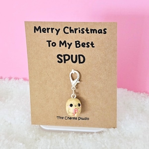May include: A brown card with the text "Merry Christmas To My Best SPUD" and a small, brown, clay charm of a potato holding a pink lollipop. The charm is attached to a silver clip.