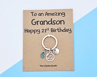 May include: A brown card with the text "To an Amazing Grandson Happy 21st Birthday" and a silver keychain with a teal charm, a silver charm with the number 21, and a silver charm with the letter L. The card has the text "Tiny Charms Studio" at the bottom.