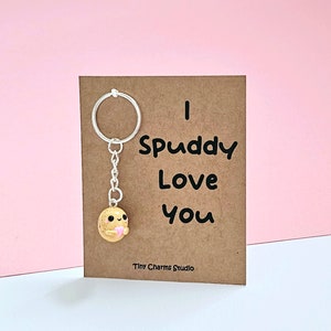 Pode incluir: Um cartão castanho com o texto "I Spuddy Love You" e um pequeno chaveiro com um amuleto de batata com um coração rosa. O texto "Tiny Charms Studio" está na parte inferior do cartão.