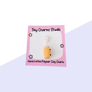 May include: A polymer clay charm in the shape of a pastry, with a silver clasp. The charm is light brown with dark brown spots, and is attached to a pink card. The card reads "Tiny Charms Studio" and "Handcrafted Polymer Clay Charm".