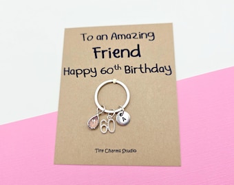 May include: A silver keychain with a pink gemstone, the number 60, and a small disc with the letter "A" hangs from a card. The card reads "To an Amazing Friend Happy 60th Birthday" and is on a pink and white background.