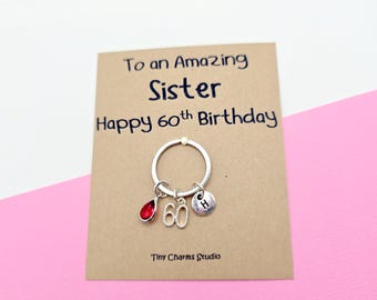 May include: A silver keychain with a red teardrop-shaped charm, the number "60", and a small disc with the letter "H". The keychain is on a card that says "To an Amazing Sister Happy 60th Birthday".