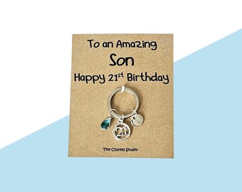 May include: A silver keychain with a teal charm, a number 21 charm, and a small initial charm. The keychain is attached to a brown card with the text "To an Amazing Son Happy 21st Birthday" and "Tiny Charms Studio". The background is light blue.