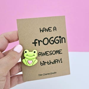 May include: A brown card with the text "Have a froggin awesome birthday!" and a green frog with a pink heart on it. The card is from Tiny Charms Studio.