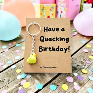 May include: A silver metal keychain with a small yellow duck charm attached to a brown card. The card features the black printed text Have a Quacking Birthday! and Tiny Charms Studio at the bottom. The duck has a small orange beak and black eyes.
