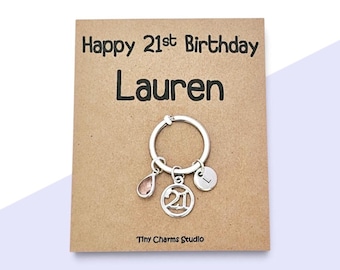 May include: A silver-toned keyring attached to a tan card printed with Happy 21st Birthday Lauren and Tiny Charms Studio. The keyring features three charms: a pink teardrop, a circular charm with the number 21, and a small disc engraved with the letter L.