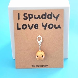 May include: A brown card with the text "I Spuddy Love You" and a small brown potato charm with a smiling face attached to a silver clip. The card is on a white surface.