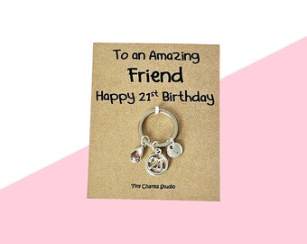 May include: A silver keychain with a charm that says "21" and a pink teardrop-shaped charm. The keychain is attached to a brown card with the text "To an Amazing Friend Happy 21st Birthday". The background is white and pink.