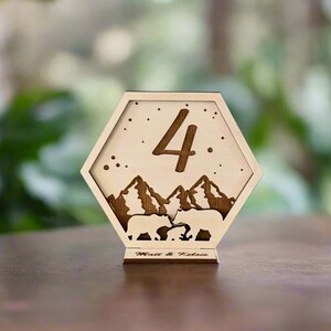 May include: Wooden table number with a hexagonal shape, featuring a mountain scene with two bears and the number "4" in the centre. The base of the table number is engraved with the text "Matt & Kelsie".