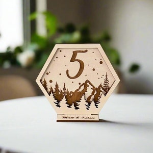 Puede incluir: Número de mesa de madera con un diseño hexagonal que presenta una escena de montaña y el número 5. La base del número de mesa está grabada con el texto "Matt & Felice".