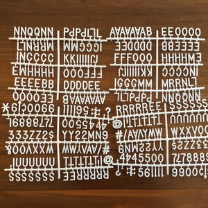 May include: White plastic letter tiles with a variety of letters, numbers, and symbols. The tiles are arranged in rows on a wooden surface.