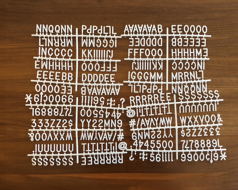 May include: White plastic letter tiles with a variety of letters, numbers, and symbols. The tiles are arranged in rows on a wooden surface.