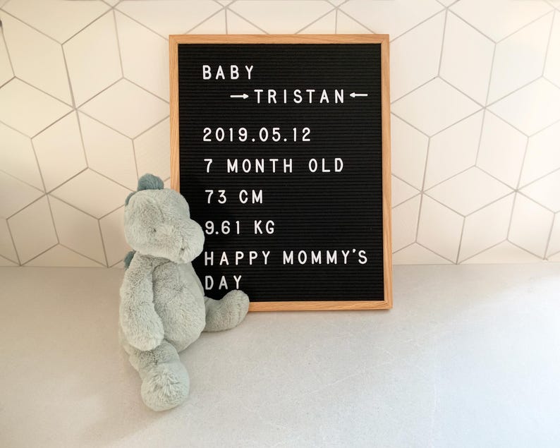 May include: A black felt letter board with white letters spelling "Baby Tristan - 2019.05.12 - 7 month old - 73 cm - 9.61 kg - Happy Mommy's Day". A green plush dinosaur toy is in the foreground.