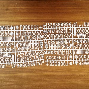 K&ouml;nnte beinhalten: Wei&szlig;e Kunststoffbuchstaben mit verschiedenen Buchstaben, Zahlen und Symbolen. Die Buchstaben sind in Reihen und Spalten angeordnet, wobei jede Kachel ein einzelnes Zeichen enth&auml;lt. Die Buchstaben befinden sich auf einer Holzoberfl&auml;che.